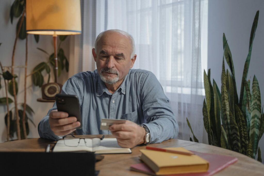 Elderly man using smartphone for online shopping at home, holding credit card near desk plants.