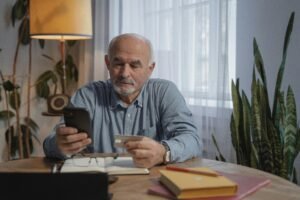 Elderly man using smartphone for online shopping at home, holding credit card near desk plants.